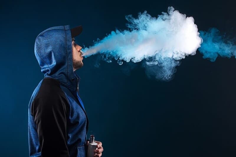 Jovem de jaqueta azul exalando nuvem após uso contínuo do vape